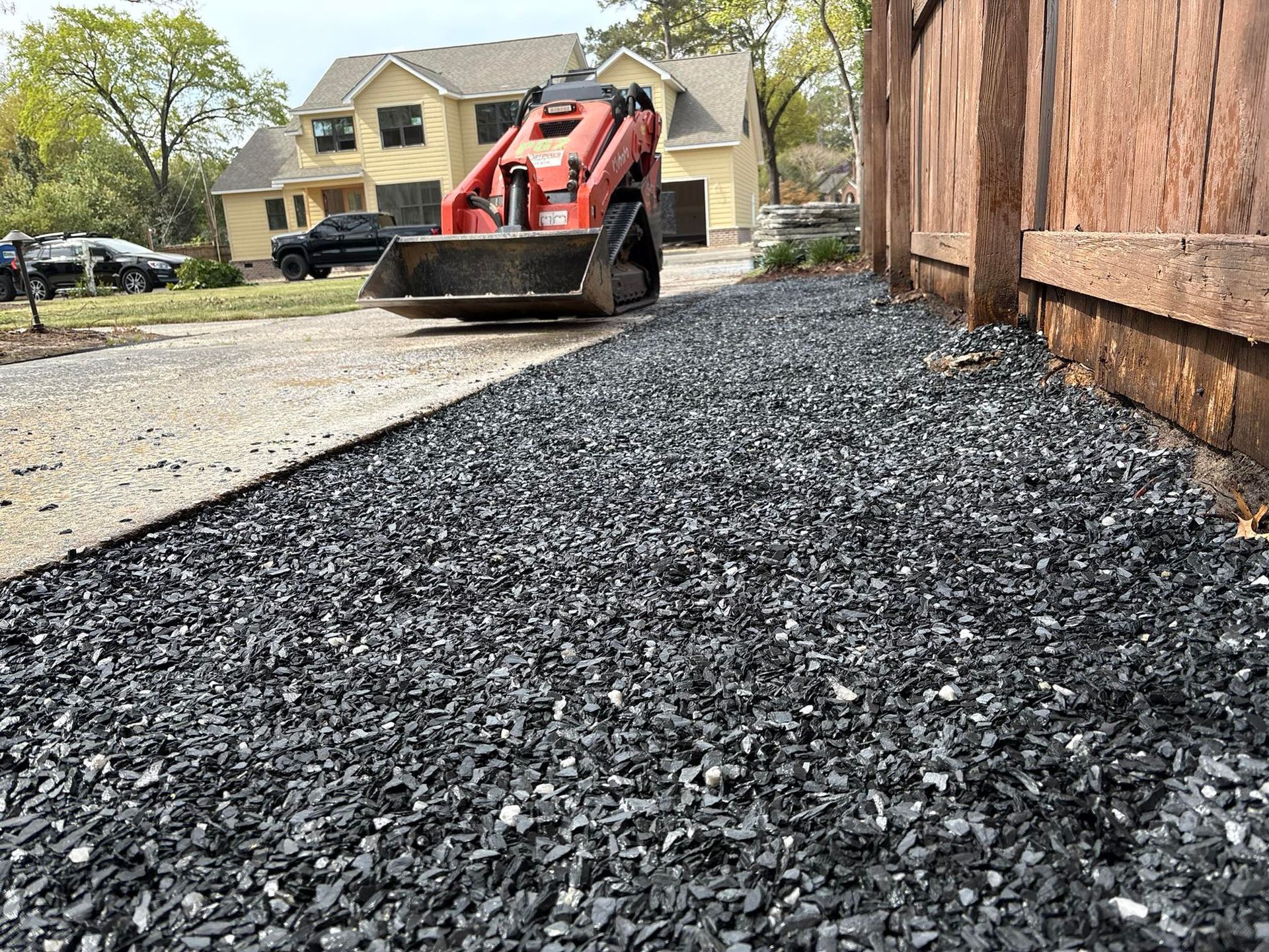 Small orange front-end loader placing dark gravel next to a wooden fence and driveway of a house.