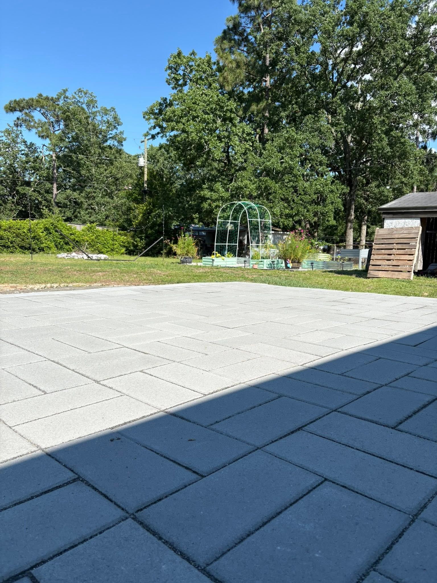Paved patio in sunlight with a grassy yard, trees, and a small structure in the background.