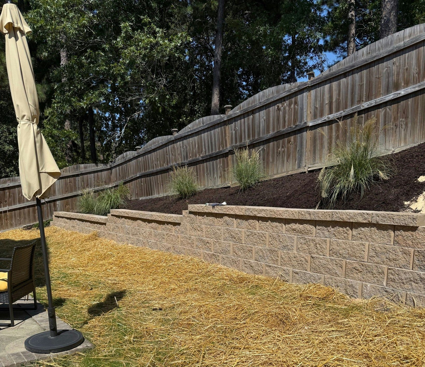 Stone retaining walls with plants and a wooden fence; a patio umbrella is on the left.