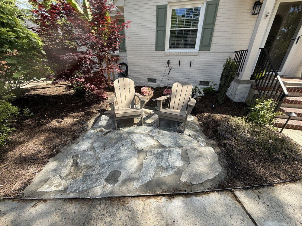 Two Adirondack chairs on a stone patio in front of a white house with a red-leaved tree.