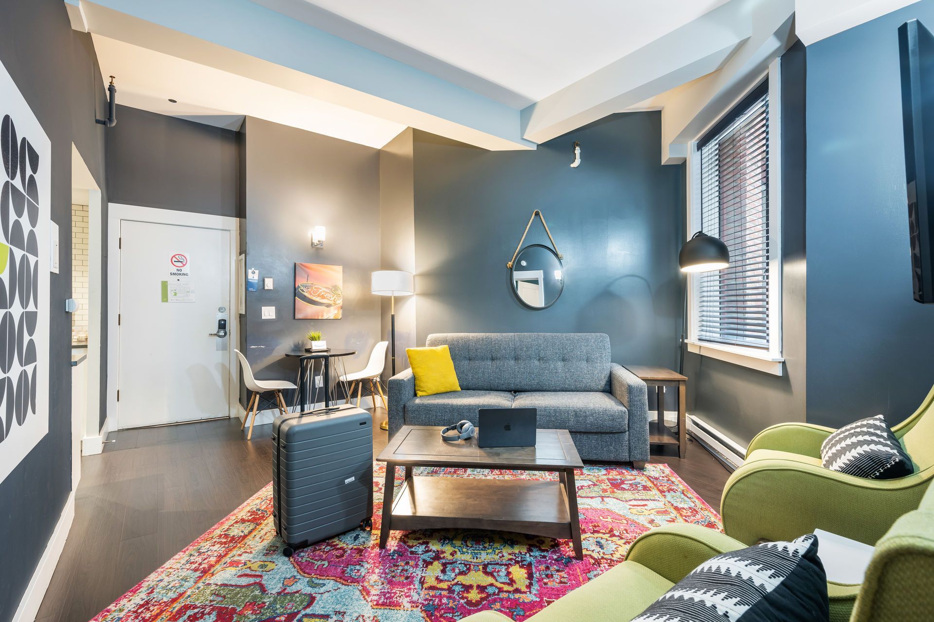 Living room with blue-gray walls, gray sofa, colorful rug, green armchairs, wooden coffee table, suitcase, and artwork.
