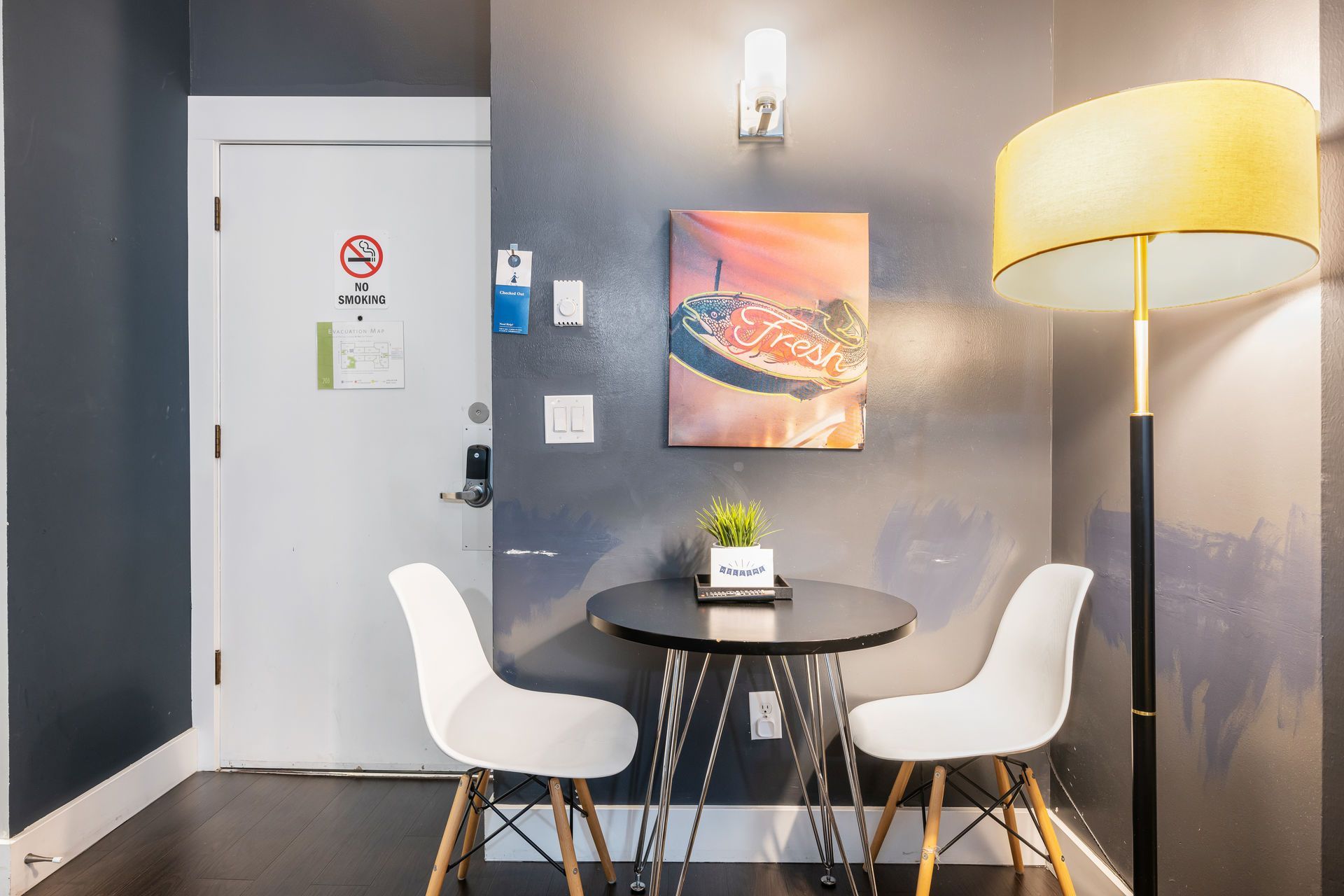 Small dining area: round black table, two white chairs, art on gray wall, standing lamp.