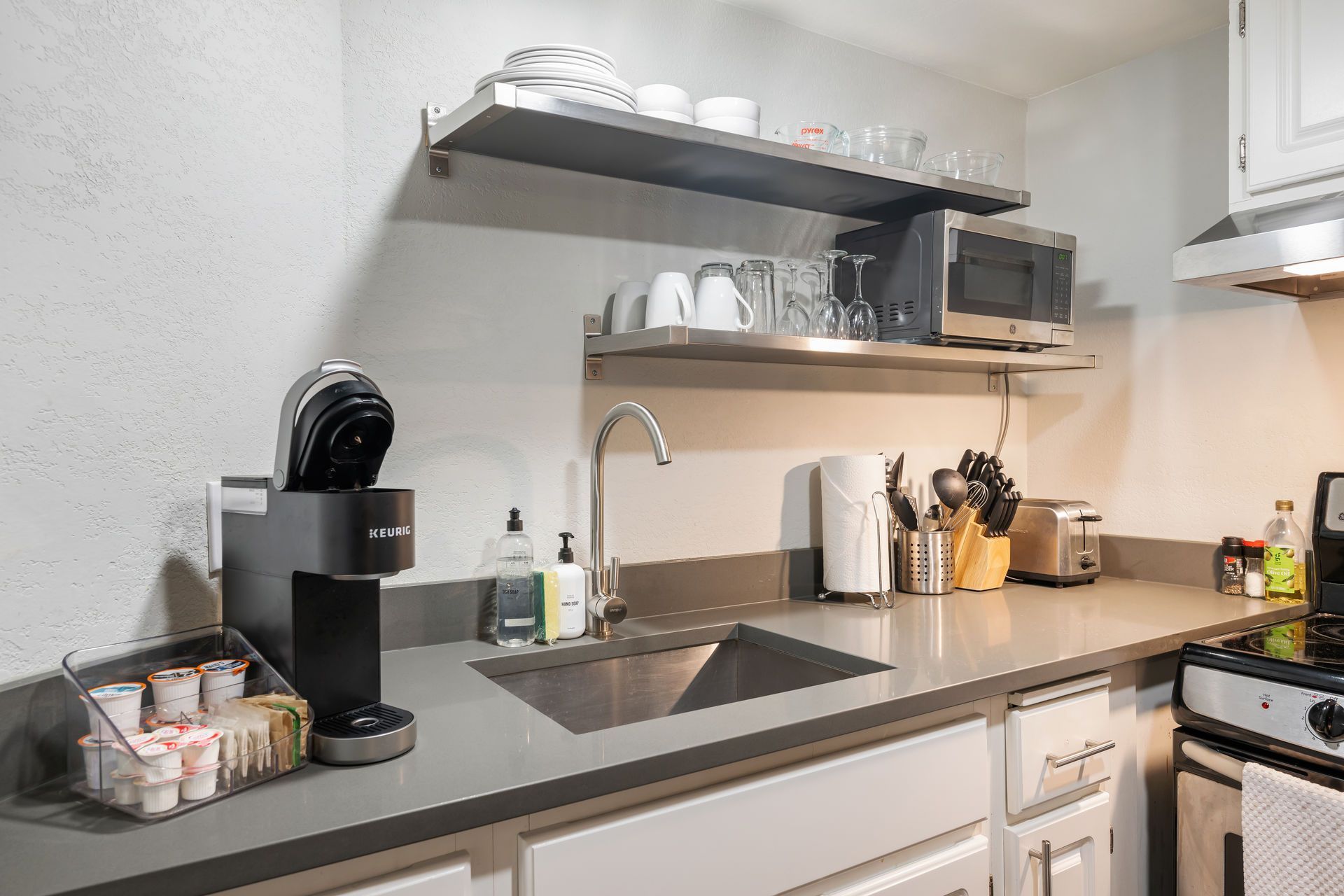 Small kitchen with stainless steel shelves, Keurig coffee maker, sink, countertop, and microwave.