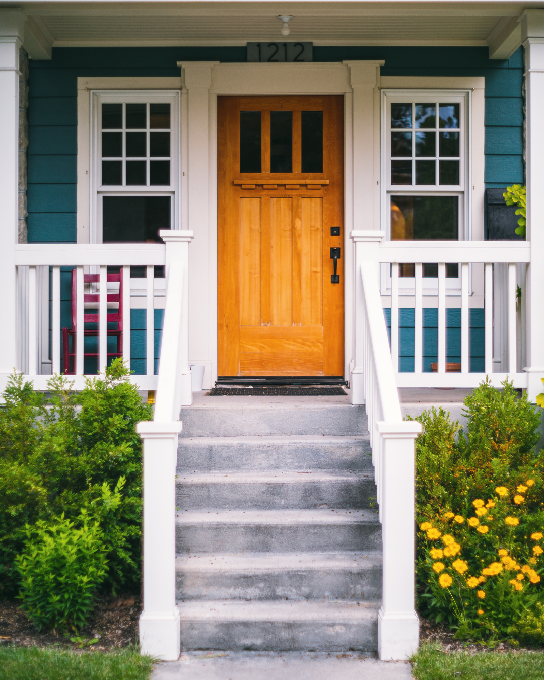 Light orange wooden door on a blue house with white trim and steps, flanked by windows and shrubs.