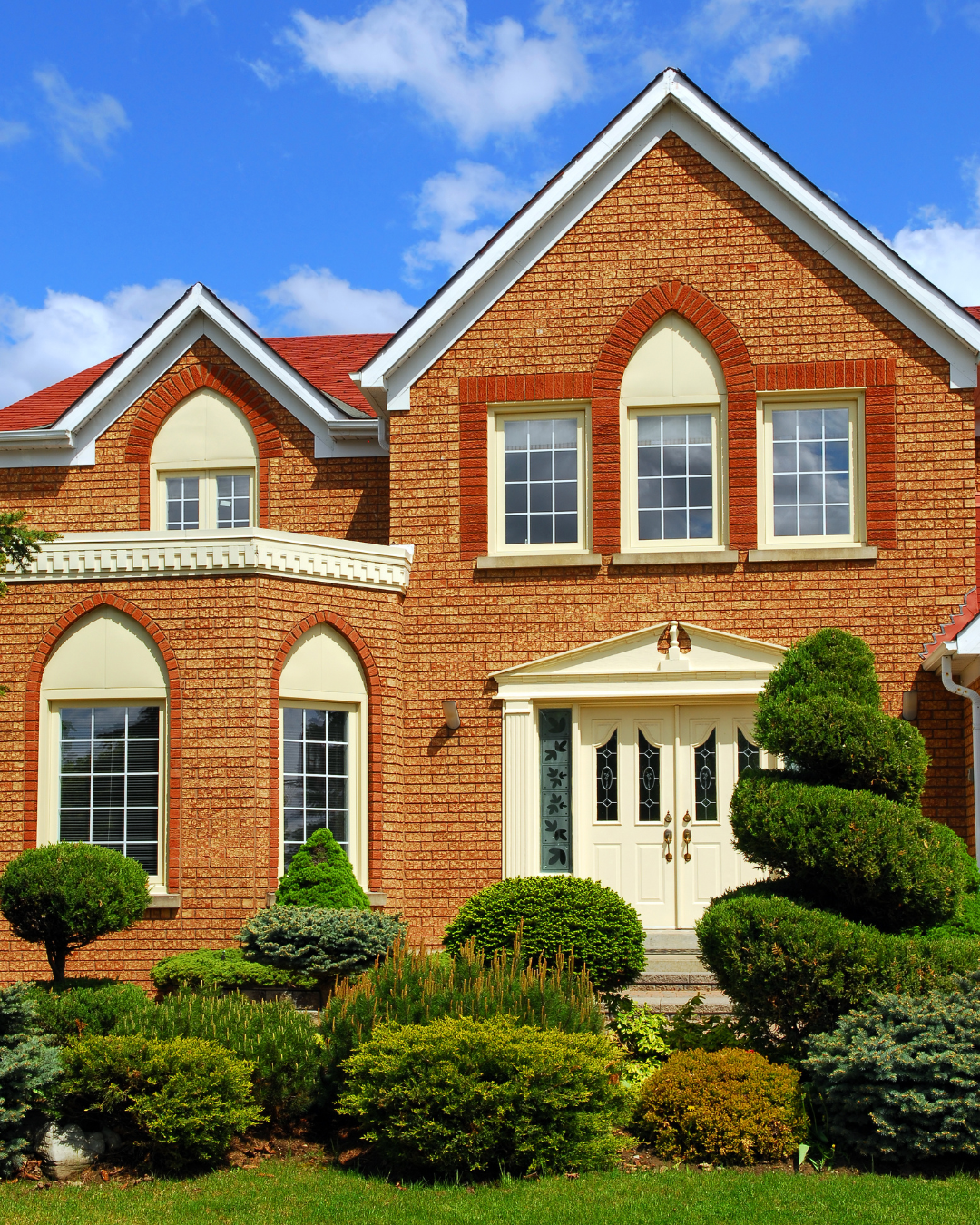 Two-story brick house with arched windows, cream trim, and lush green landscaping against a blue sky.