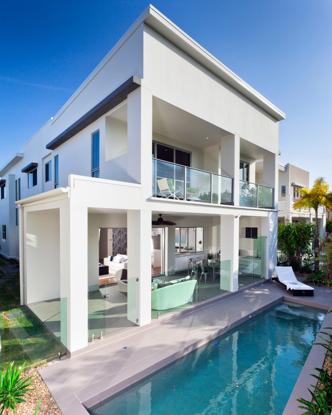 Two-story white house with a pool. Balcony, glass railings, and patio. Sunny day.