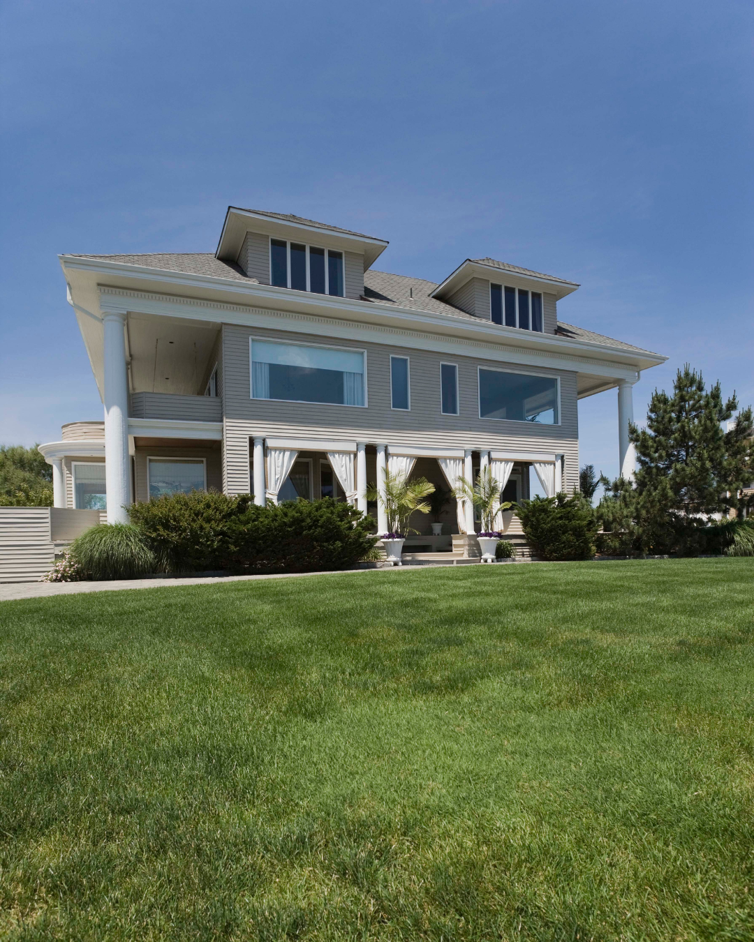 Two-story house with large windows, white pillars, and a green lawn under a blue sky.