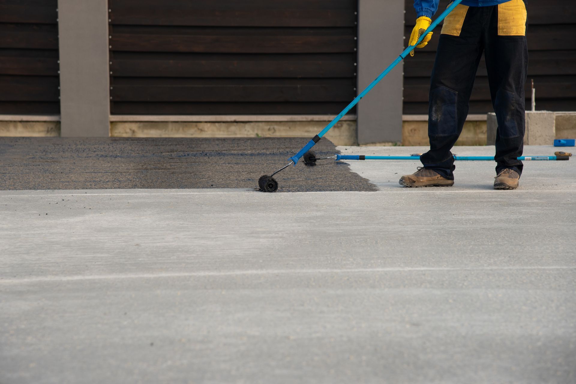 Worker rolls black coating onto concrete near fence in construction zone.