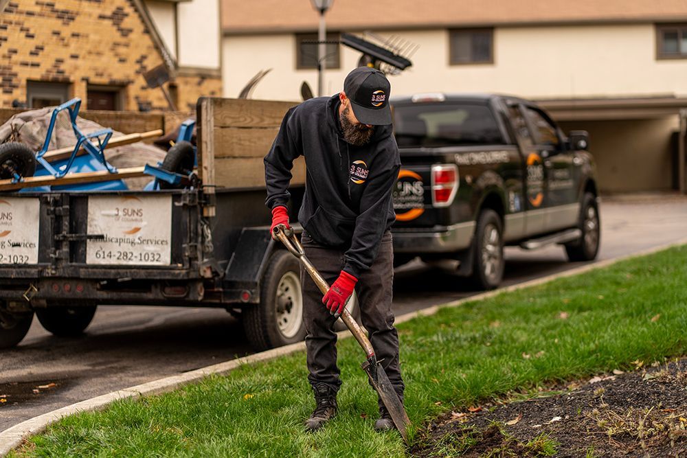 Man digging in lawn near a trailer and truck, wearing black hoodie and red gloves.