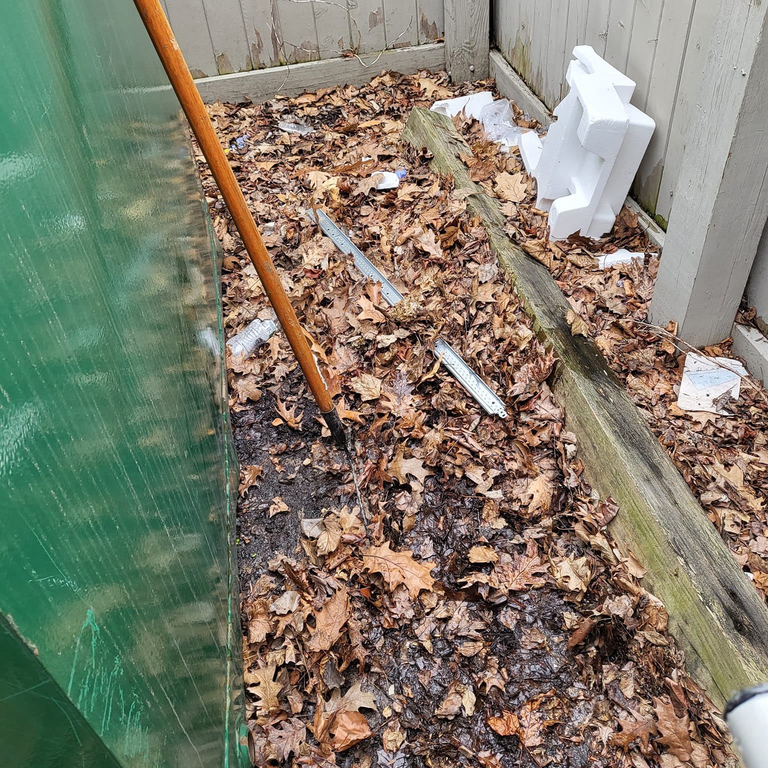 A person rakes leaves from a damp garden bed against a wooden fence.