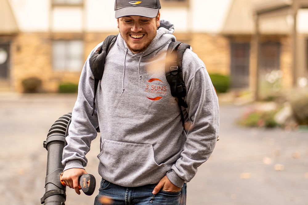 Man in gray hoodie using leaf blower; smiles outdoors.