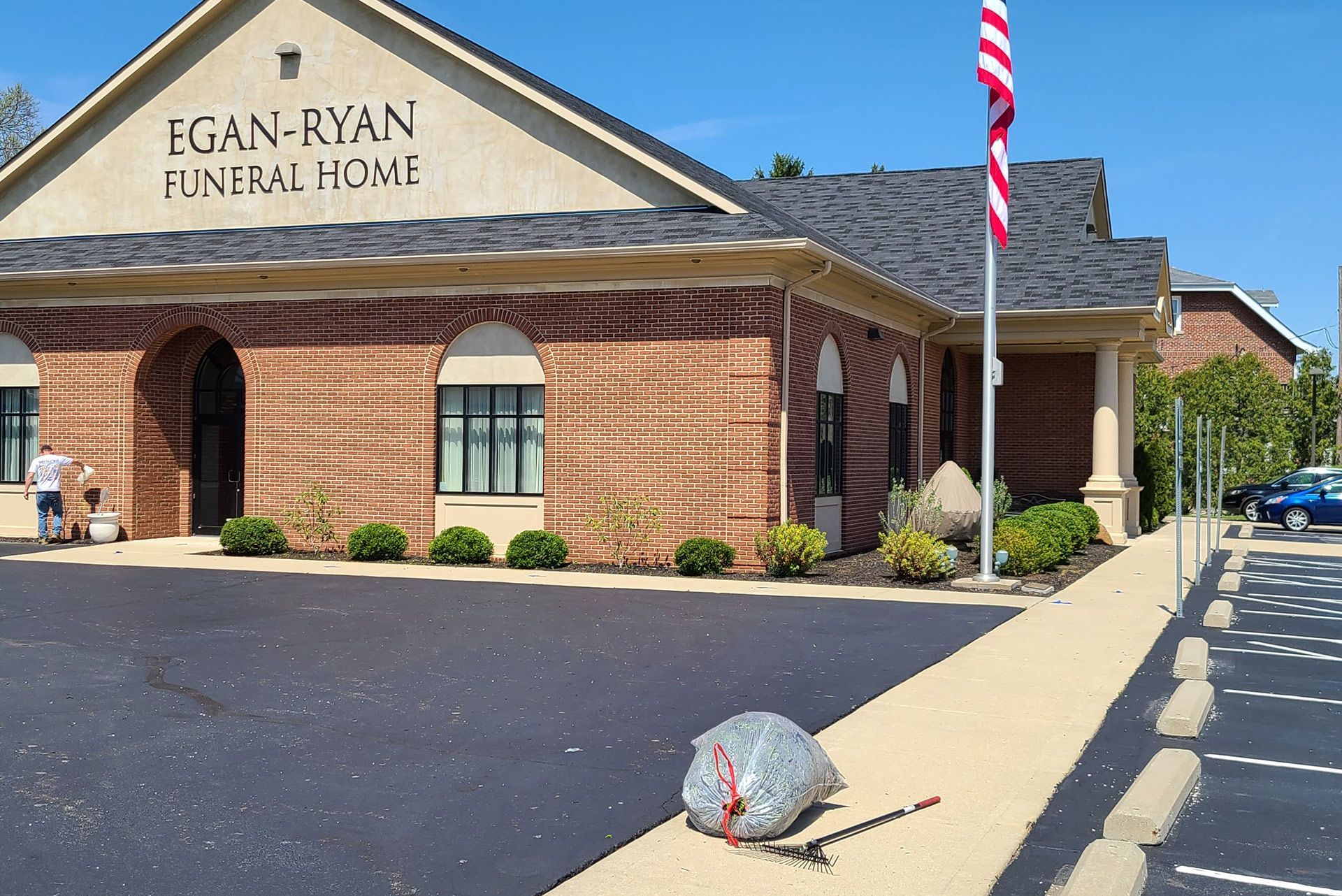 Exterior of Egan-Ryan Funeral Home, red brick building with American flag. Person working near door, trash bag on sidewalk.
