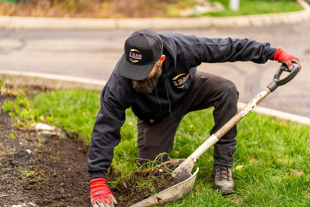 A man kneels in a garden, digging with a shovel. He wears a black hat, jacket, and red gloves.