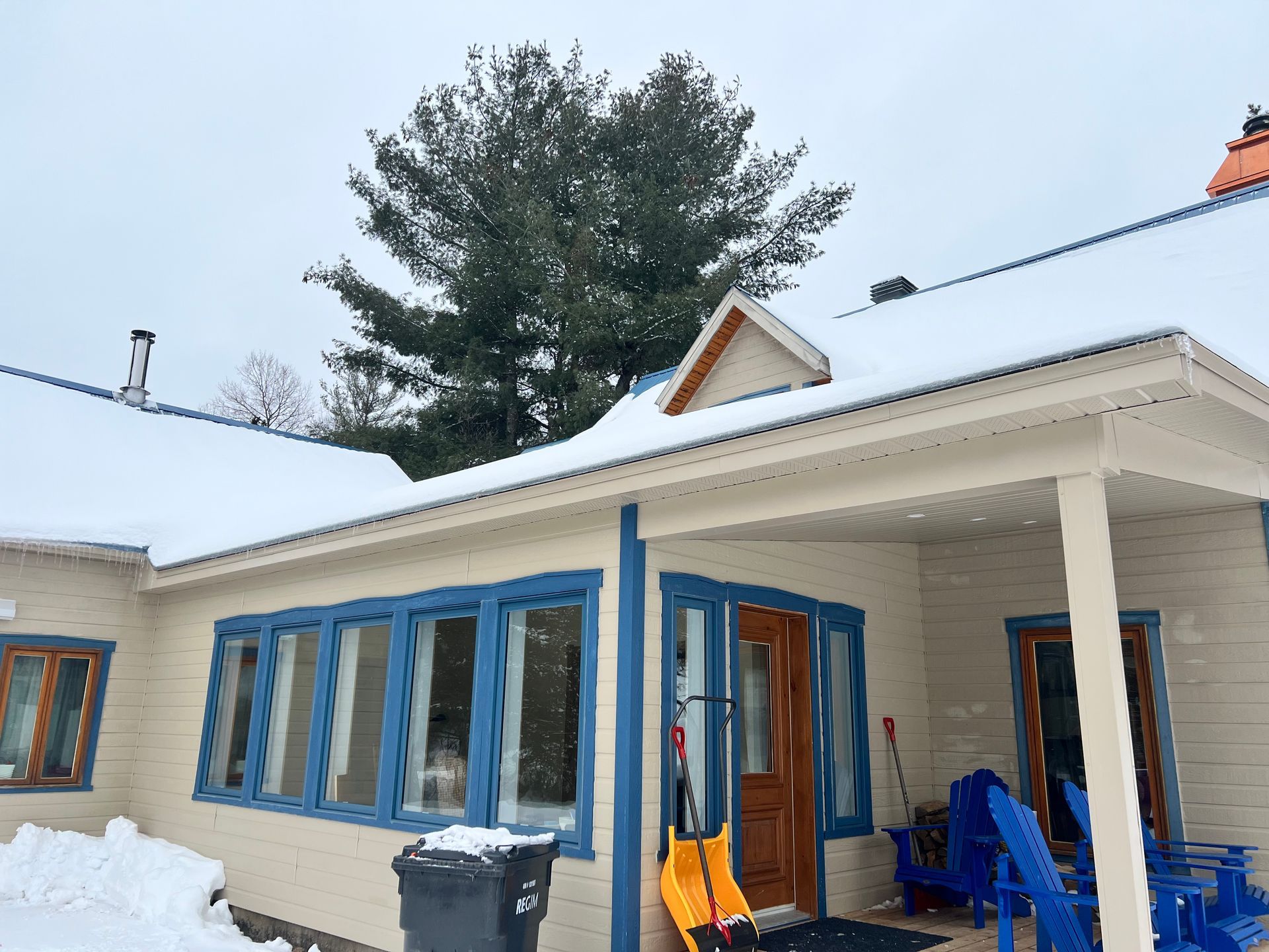 Une maison aux tons clairs, avec des encadrements de fenêtres bleus, une porte en bois et un porche, le tout recouvert d'une légère neige sous un ciel d'hiver.