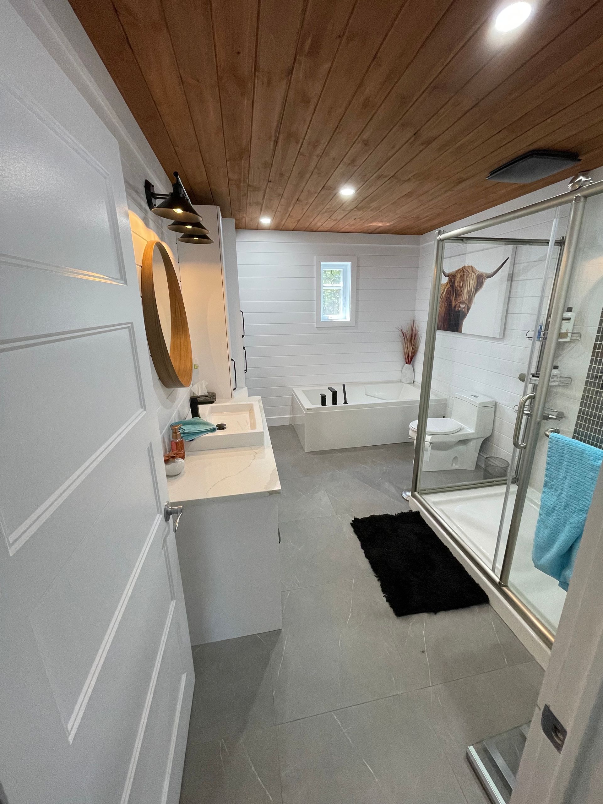 Une salle de bains moderne comprend un plafond lambrissé, des murs blancs, un meuble-lavabo avec un miroir rond, une baignoire et une douche.