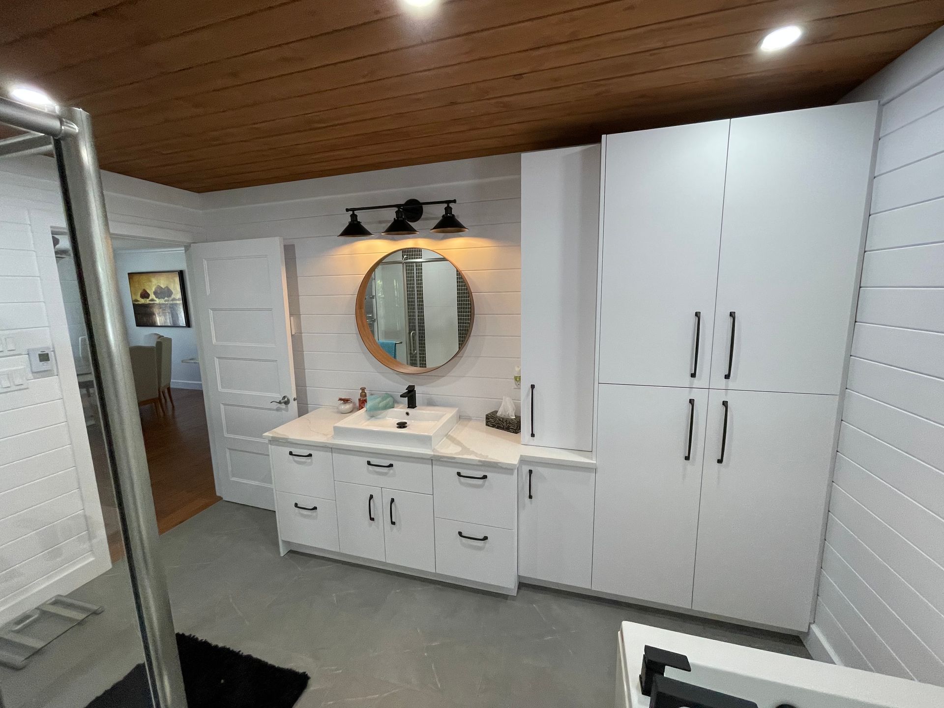 Une salle de bains lumineuse avec un meuble-lavabo blanc surmonté d'un miroir rond, une armoire de rangement haute et des murs lambrissés blancs.
