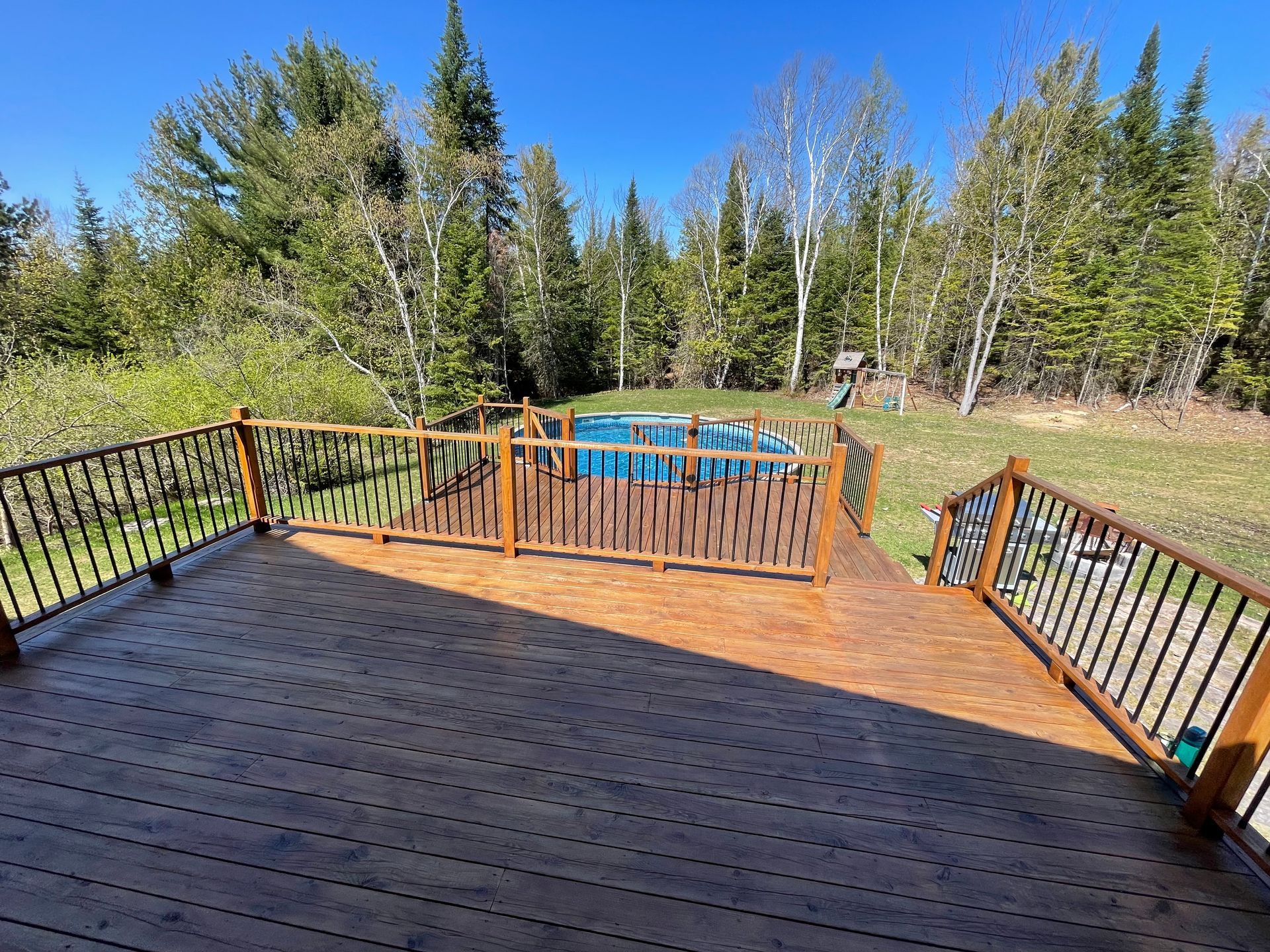 Vue depuis une terrasse en bois donnant sur une piscine dans un jardin entouré d'une dense forêt de conifères.