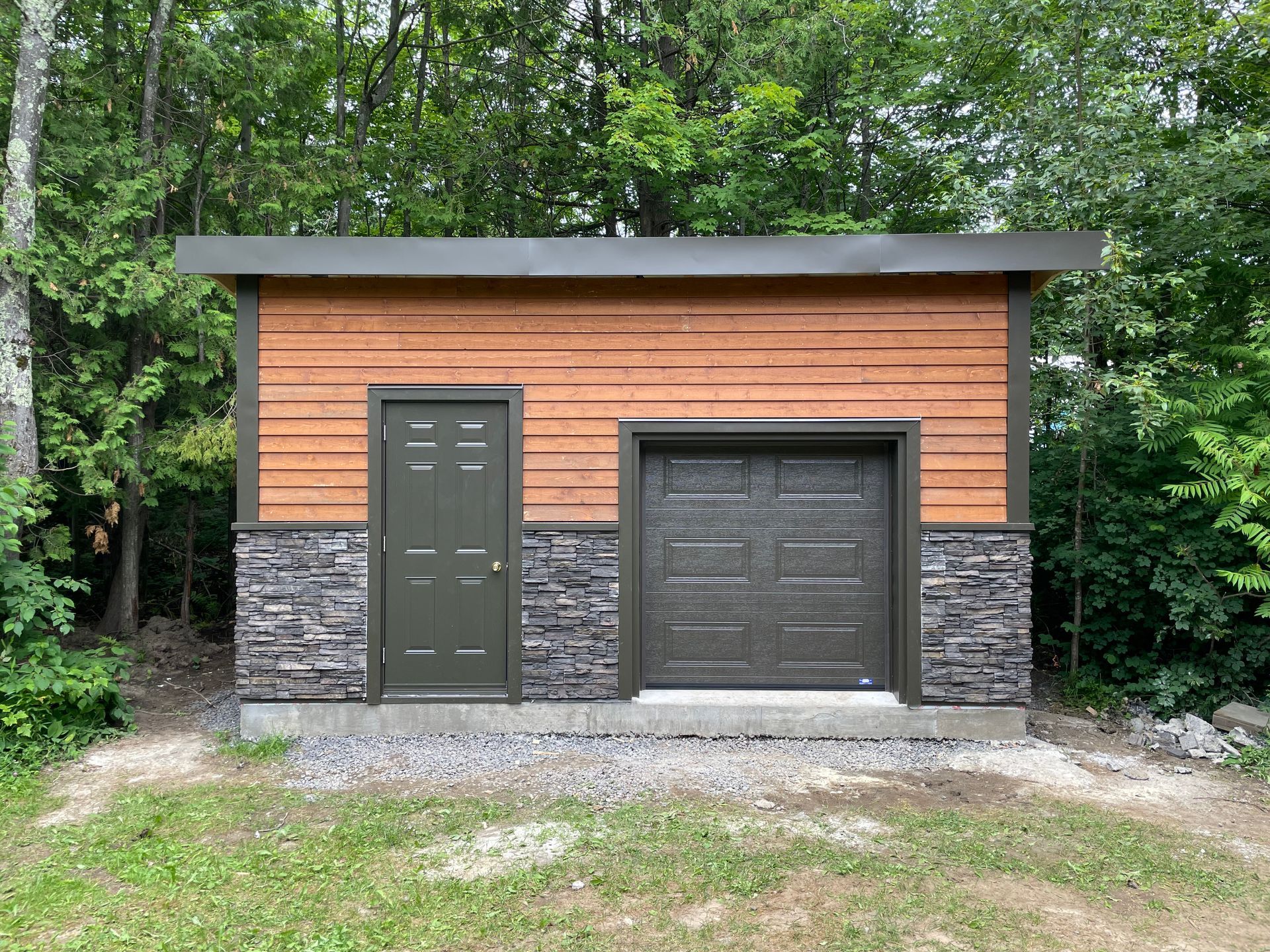 Petit cabanon avec bardage en bois et soubassement en pierre, doté d'une porte en olivier et d'une porte de garage, le tout entouré d'une forêt.