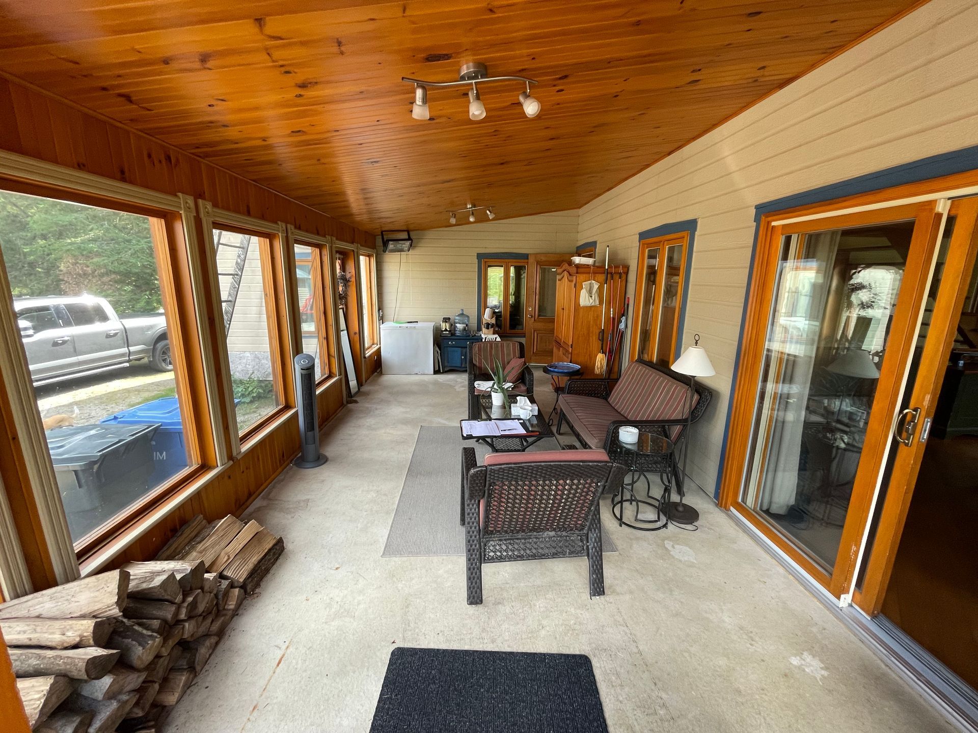 Une terrasse couverte avec un plafond lambrissé, de grandes fenêtres donnant sur un camion, des meubles en osier et un tas de bois de chauffage.