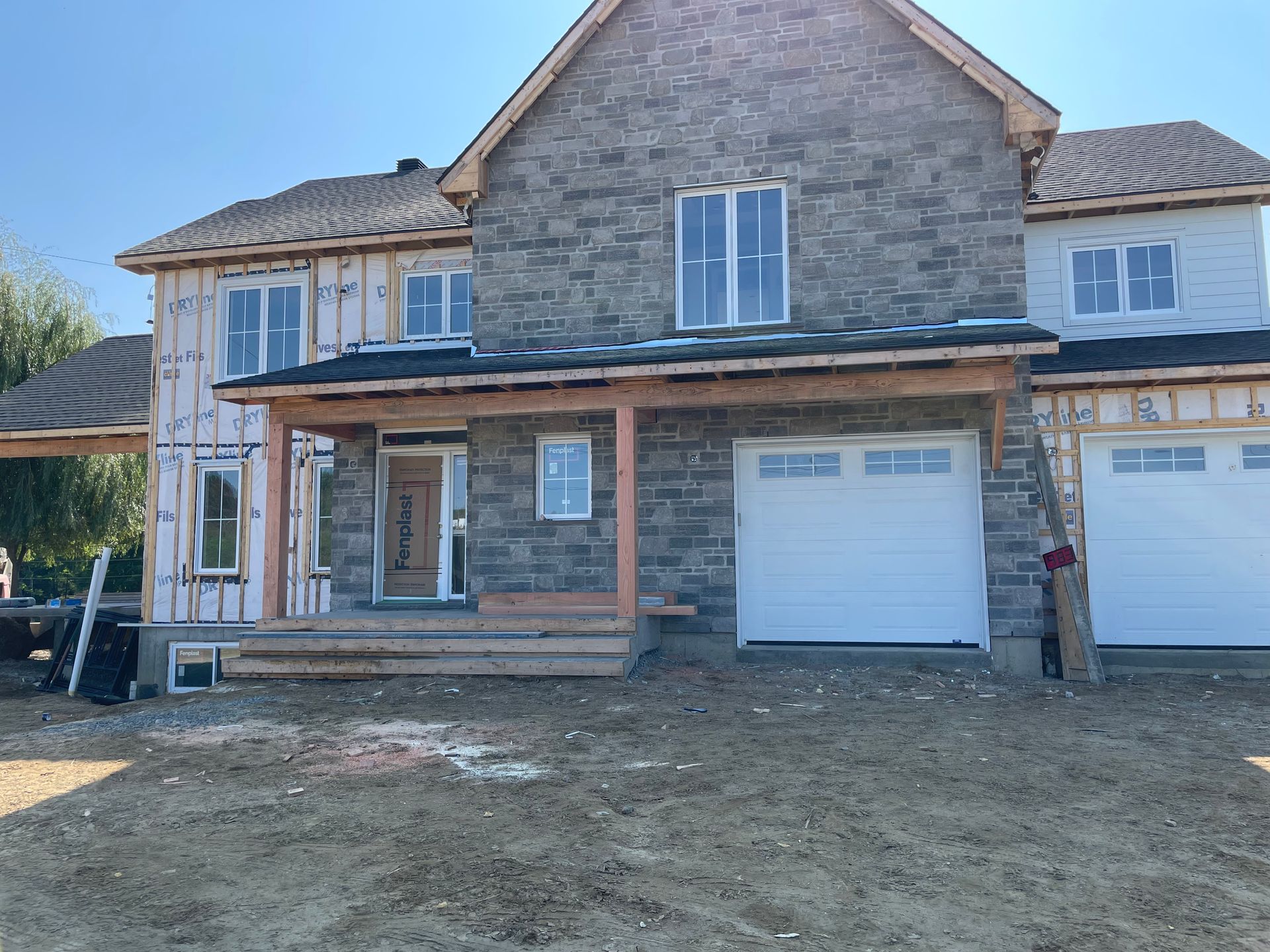 Une maison à deux étages en construction, avec un parement en pierre grise, des portes de garage blanches et un bardage inachevé, sur un terrain en terre battue.