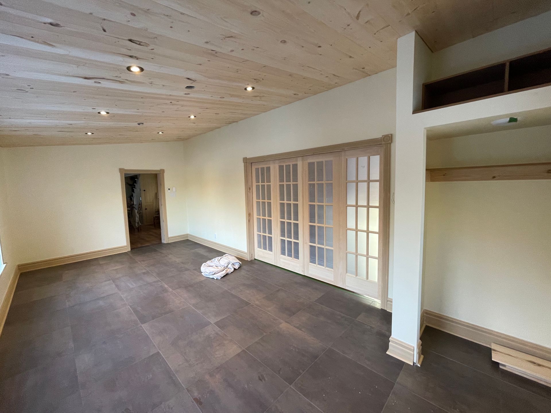 Une pièce lumineuse avec un plafond en bois clair, un sol carrelé foncé, une porte et des portes doubles vitrées.