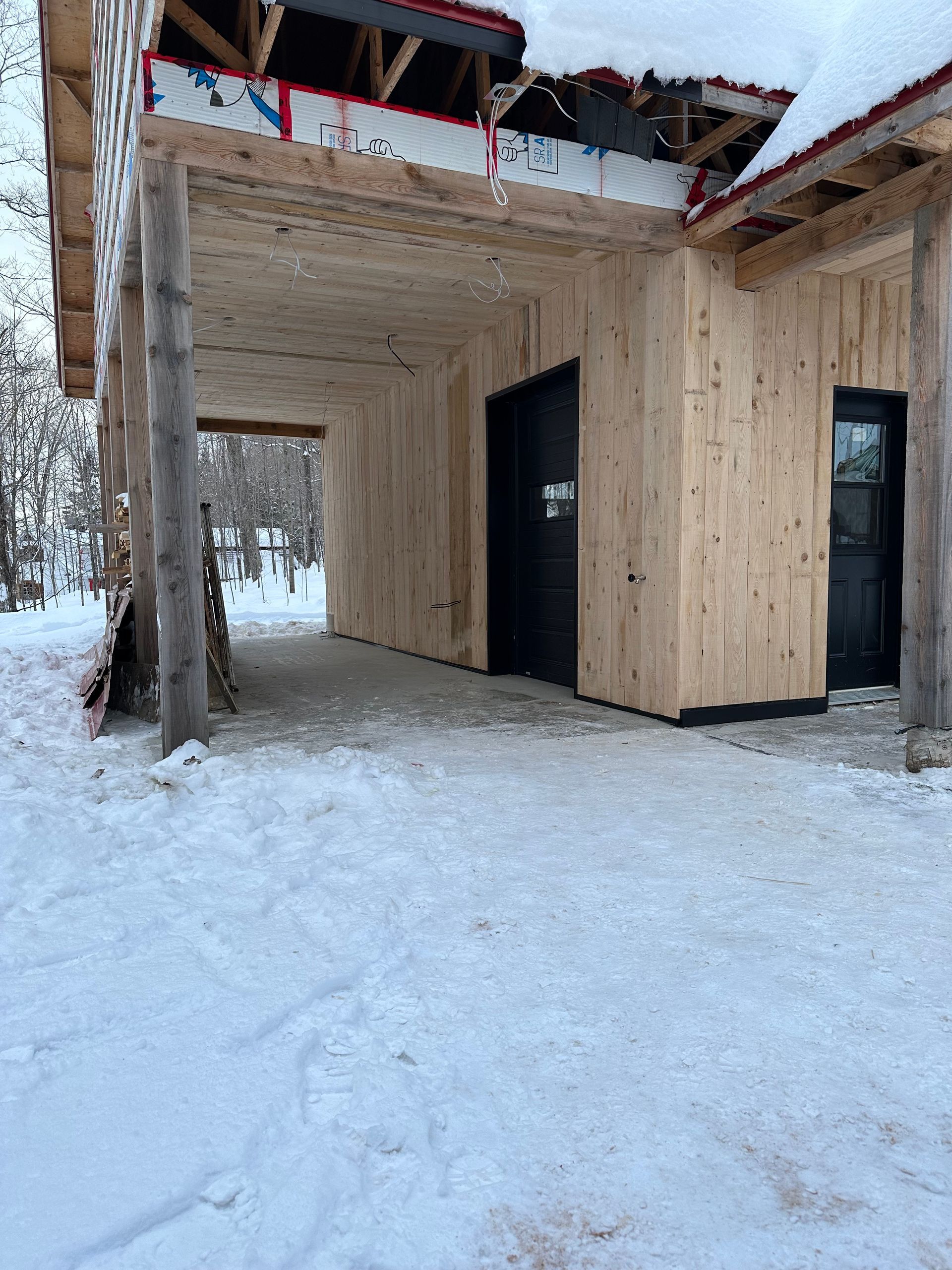 Un bâtiment en bois en construction, avec un sol enneigé et deux portes sombres, dans un décor de forêt enneigée.