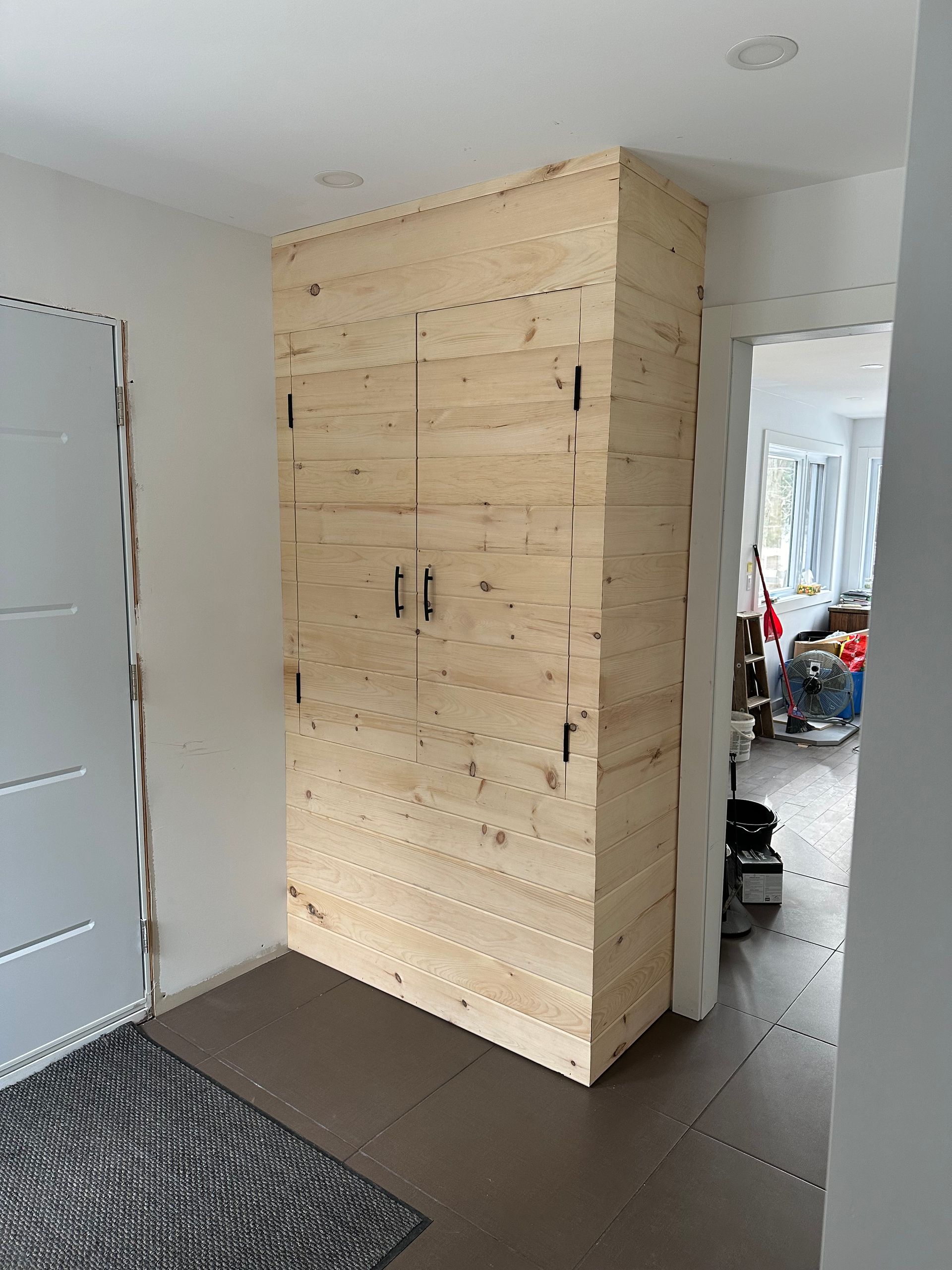 Un meuble de rangement haut, en panneaux de bois clair, encastré dans un coin de l'entrée d'une maison, situé à côté d'une porte.