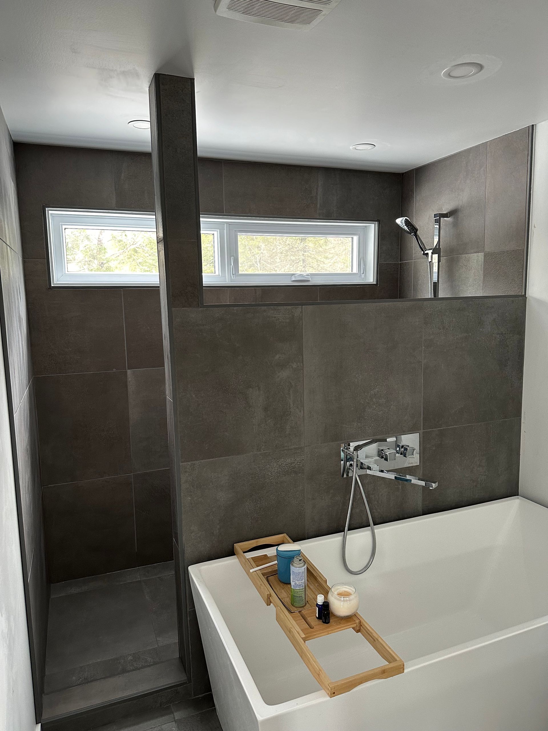 Une salle de bains moderne avec une baignoire blanche surmontée d'un plateau en bambou, jouxtant une douche à l'italienne aux murs carrelés gris foncé.