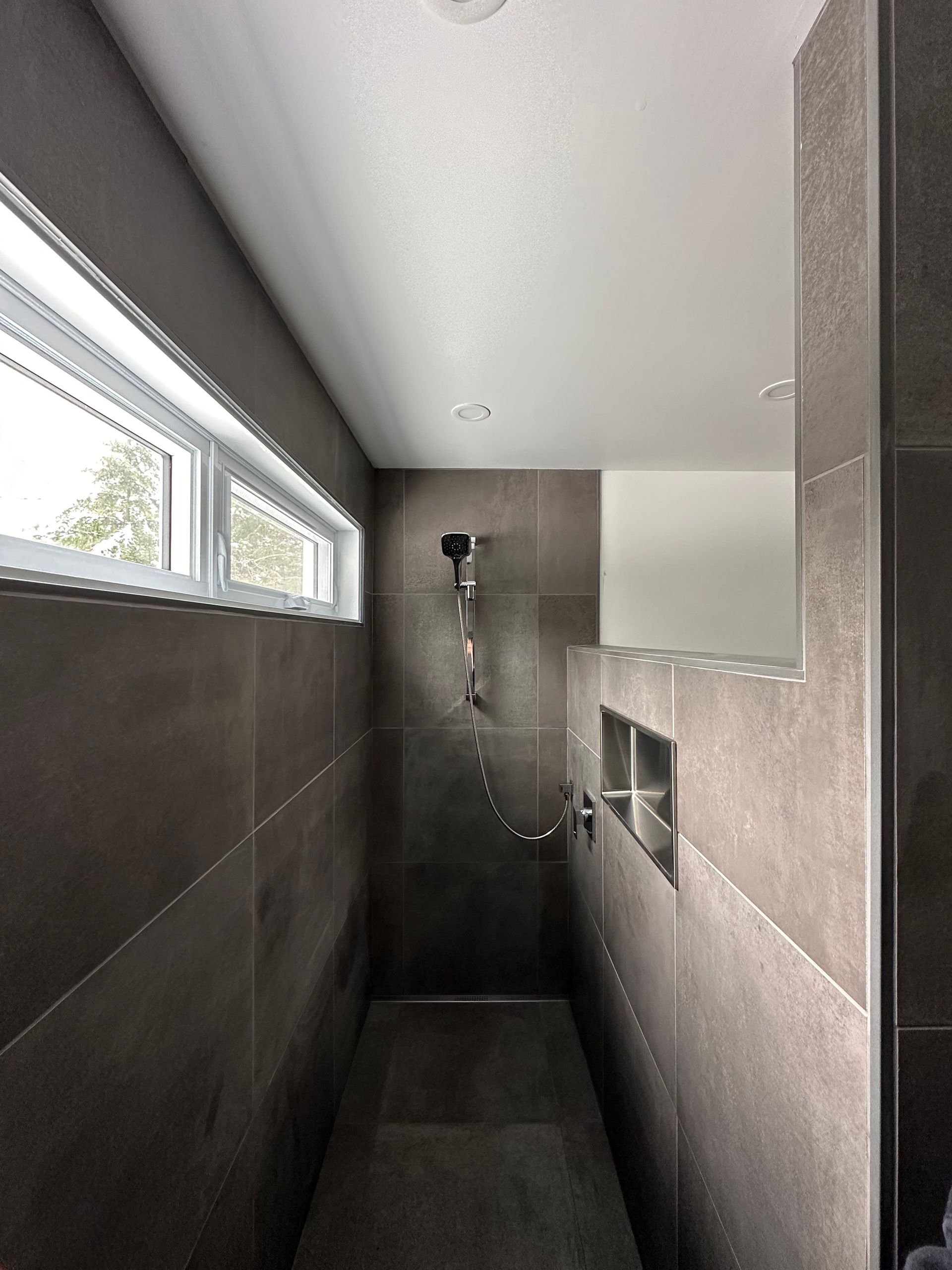 Une douche à l'italienne avec des carreaux gris foncé imitation pierre, une douchette et une fenêtre rectangulaire étroite.