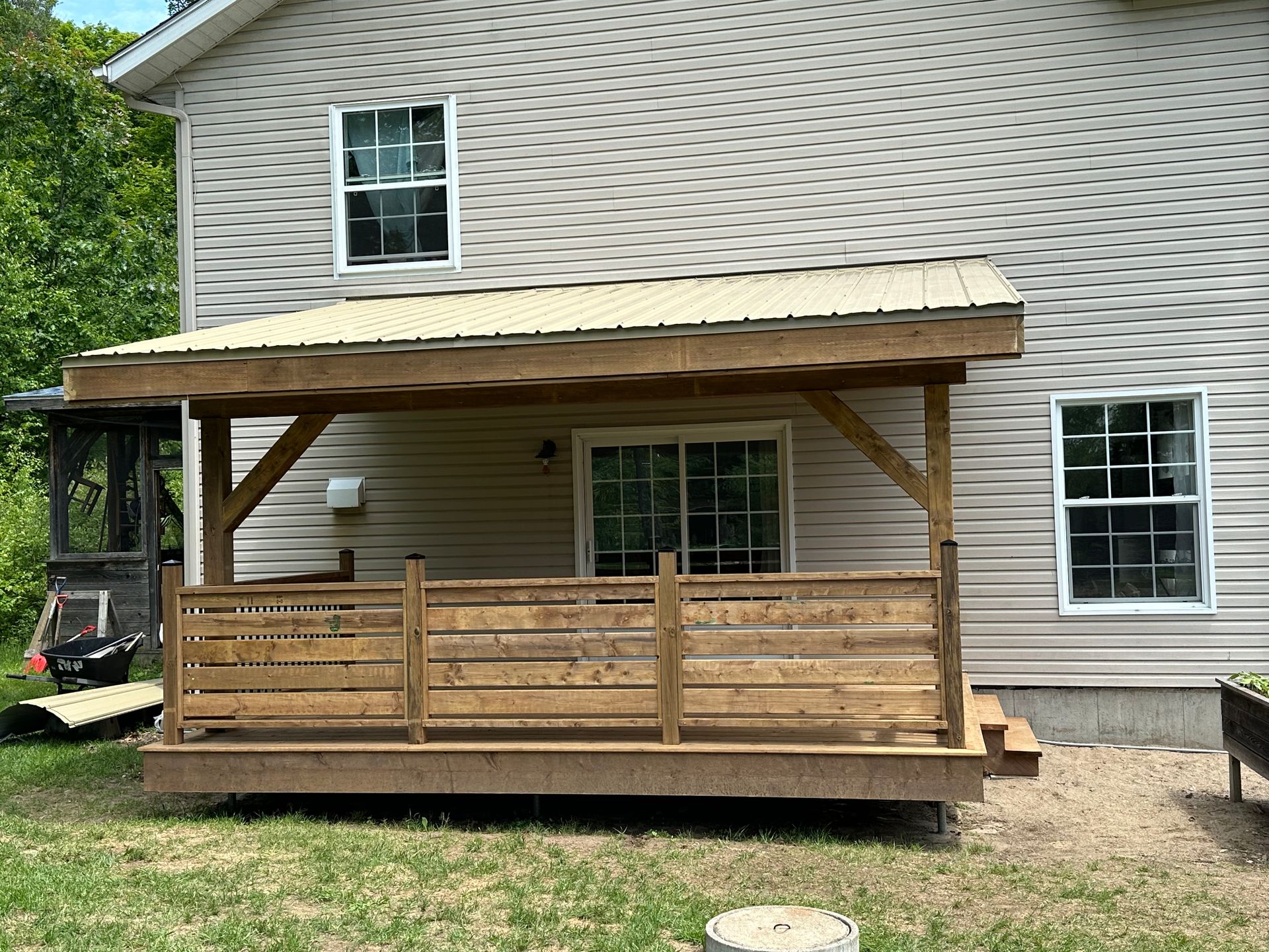 Une terrasse en bois avec un porche au toit métallique, adossée à l'arrière d'une maison au bardage horizontal de couleur claire.