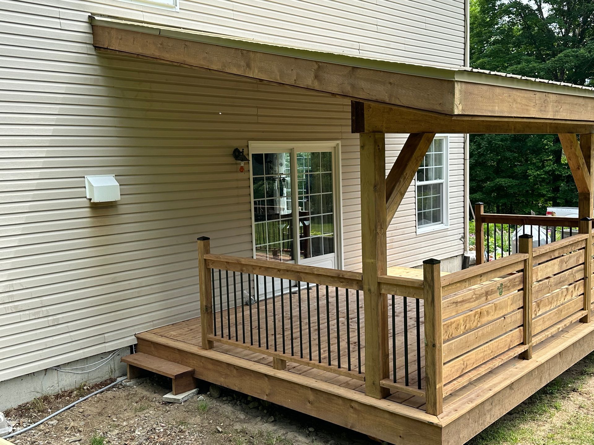 Une terrasse en bois avec un toit couvert, des rambardes noires et des lattes horizontales fixées sur le côté d'une maison aux murs beiges.