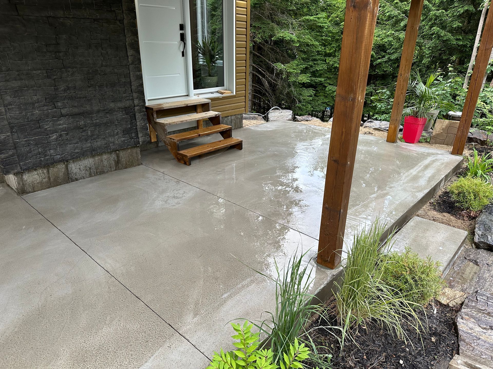 Un porche en béton humide avec des marches en bois menant à une porte blanche, encadrée par des piliers en bois et un aménagement paysager.