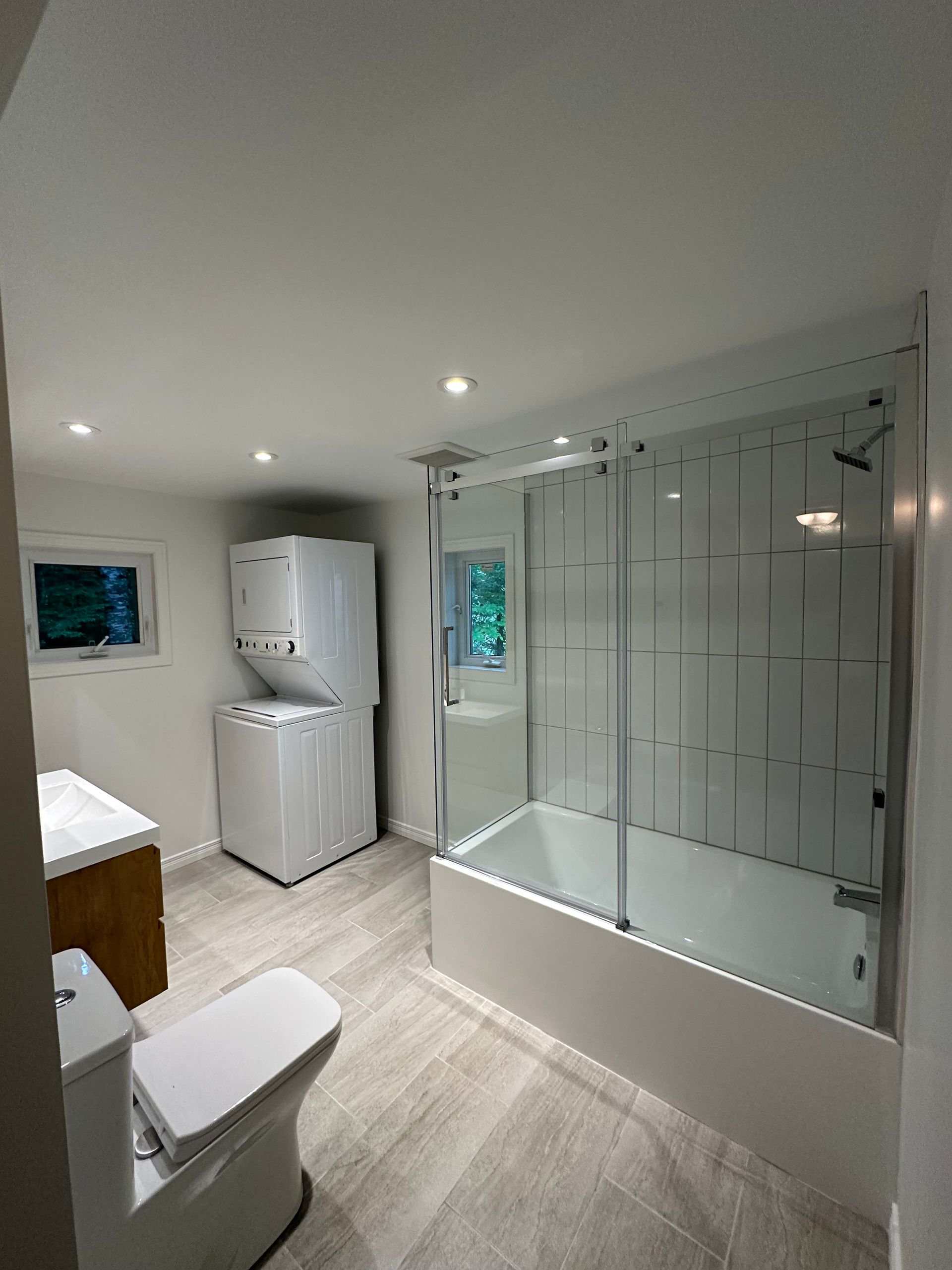 Une salle de bains comprenant une baignoire-douche combinée avec portes en verre, un lave-linge et un sèche-linge superposés, un meuble-lavabo et des toilettes.