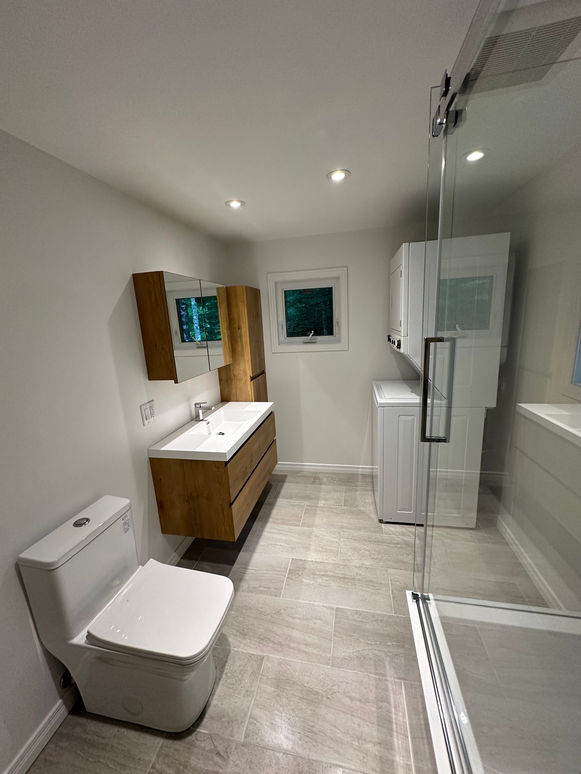 Une salle de bains moderne comprenant des toilettes blanches, un meuble-lavabo en bois avec double vasque, un lave-linge séchant superposé et une douche en verre.