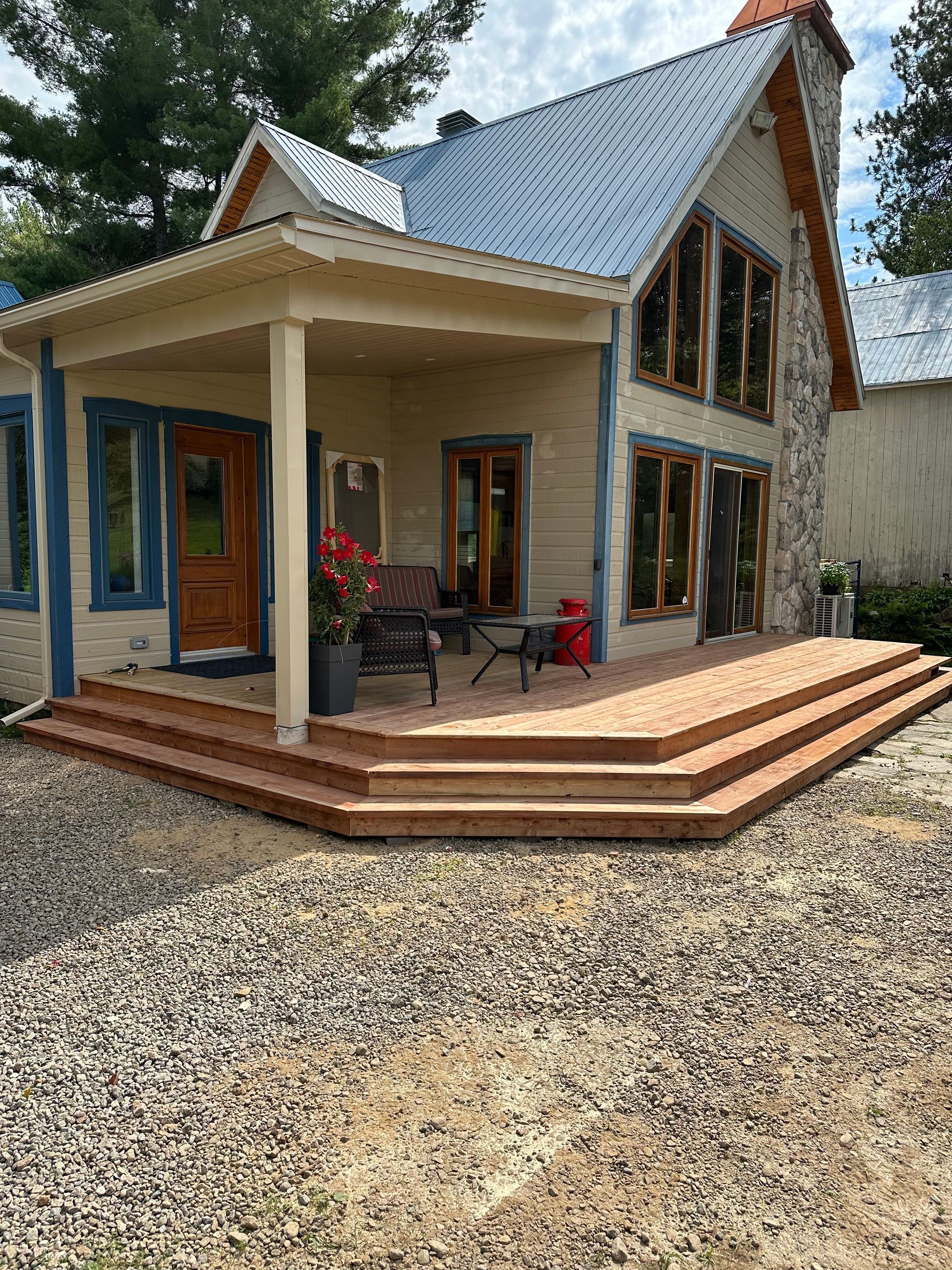 Un cottage beige avec une cheminée en pierre, des fenêtres à encadrement bleu et une large terrasse en bois à plusieurs niveaux dans une cour gravillonnée.