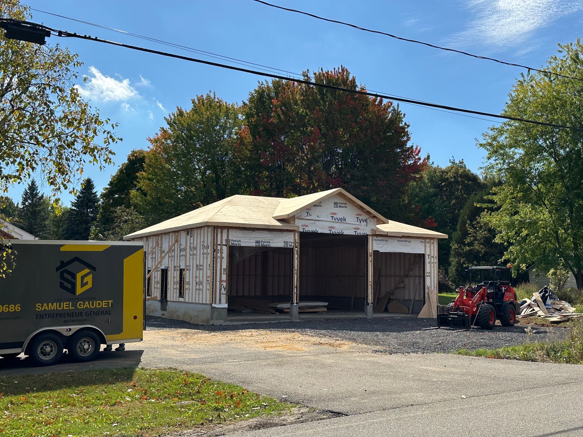 Un chantier de construction avec la structure d'un nouveau garage, une remorque de marque « SG » et un tracteur rouge sous un ciel ensoleillé bordé d'arbres.