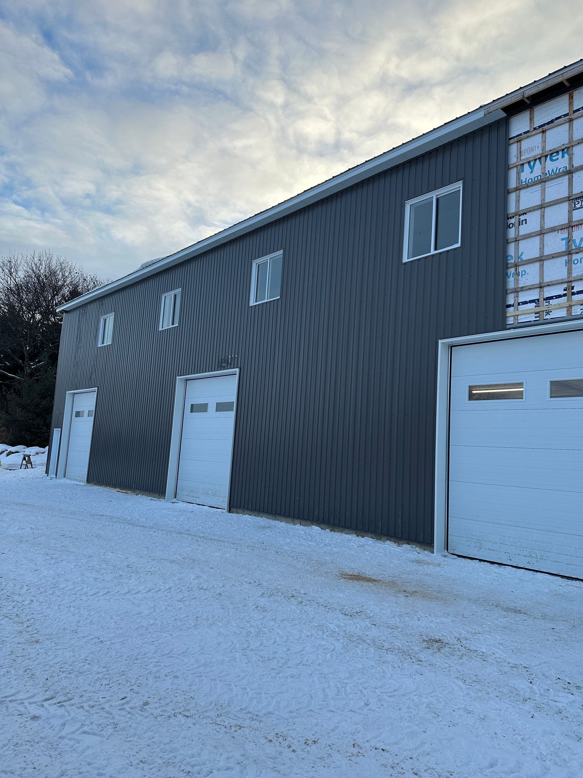 Un bâtiment aux parois métalliques grises, avec trois portes de garage blanches et des fenêtres à l'étage, se détachant sur un paysage enneigé.