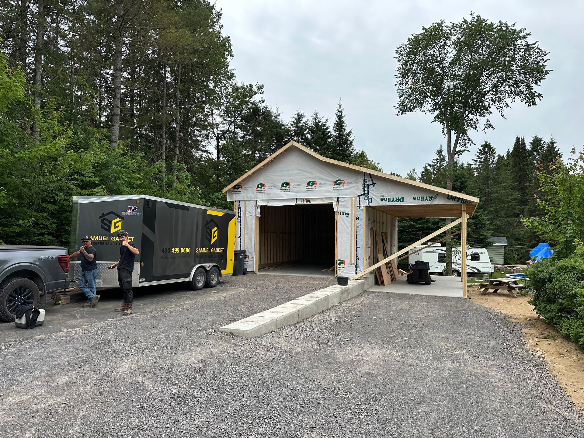 Deux personnes se tiennent à côté d'une caravane sombre, près d'un garage en construction avec une allée en béton, sur un terrain boisé.
