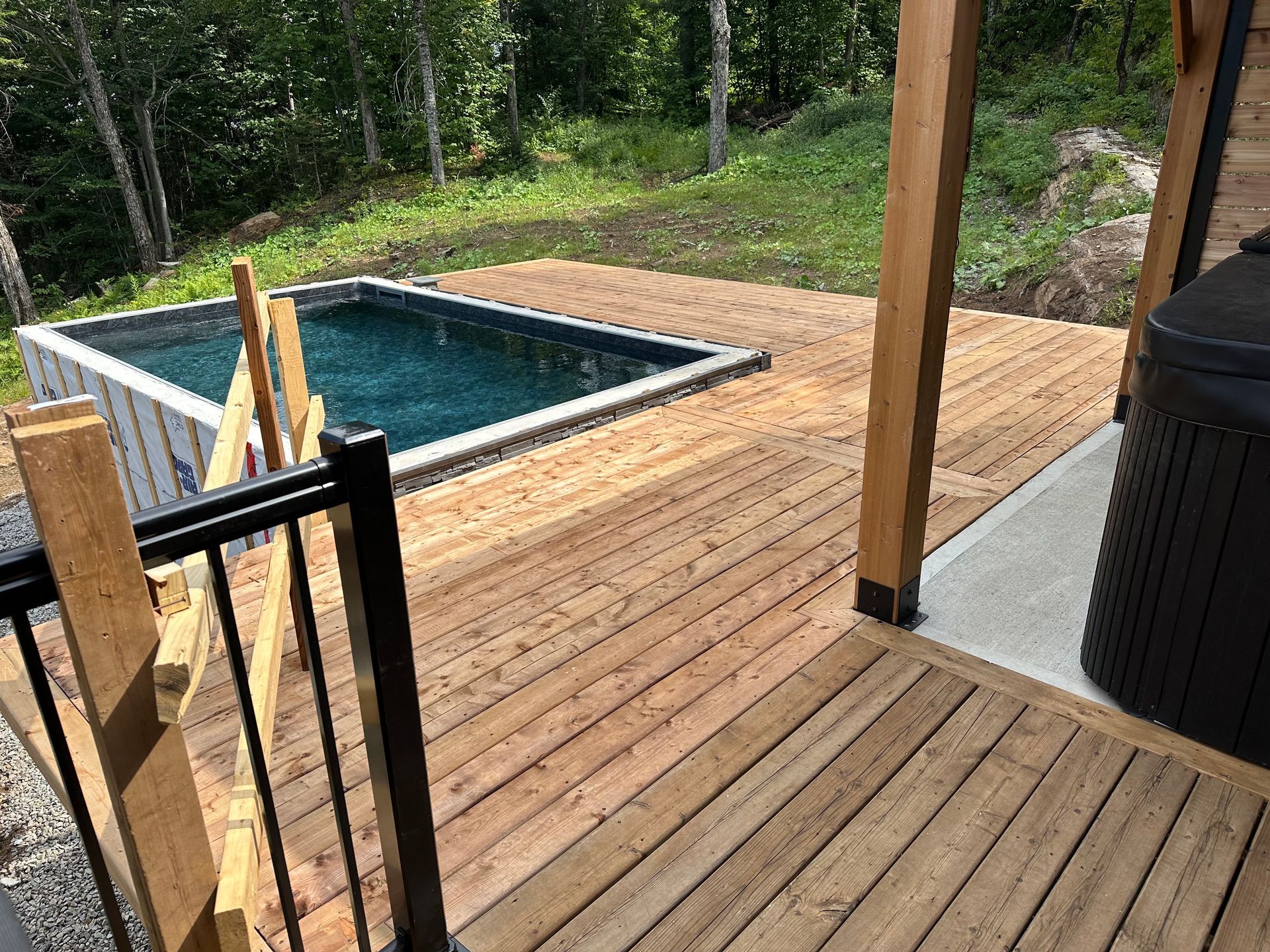 Un bassin rectangulaire aménagé sur une terrasse en bois surplombant une colline boisée, avec un jacuzzi noir visible sur la droite.