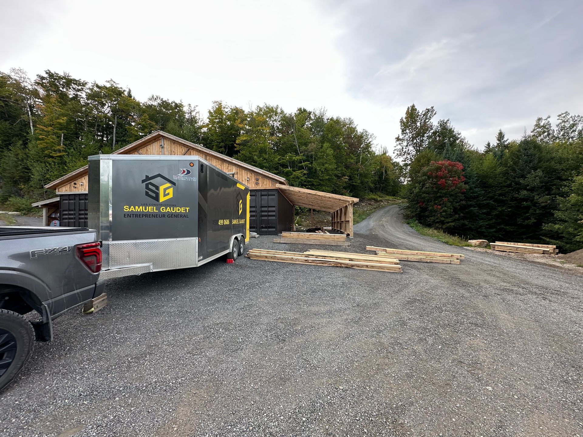Une remorque grise attelée à une camionnette stationnée sur une allée de gravier près d'un bâtiment en bois en construction.