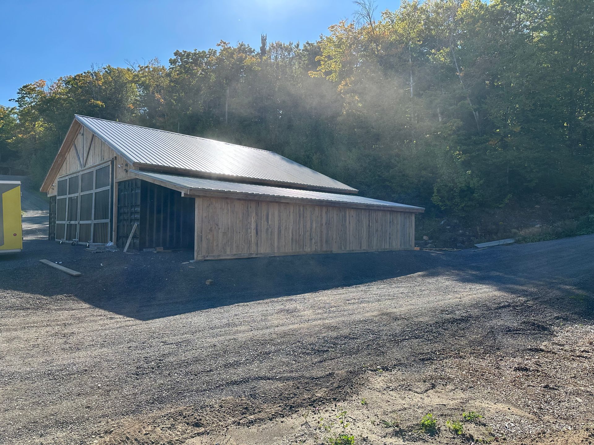 Une grange en bois avec un toit en métal en construction, située en bordure d'un terrain gravillonné, au pied d'une colline boisée.