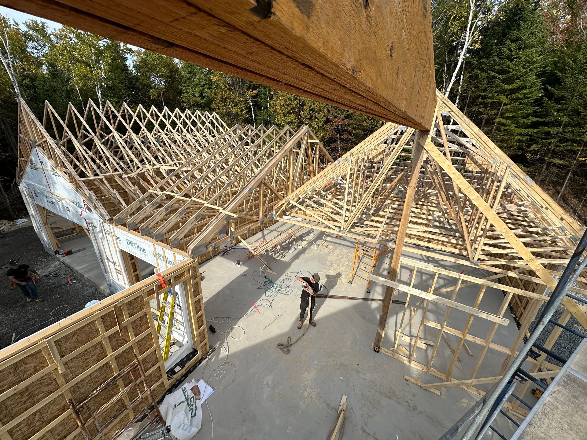 Chantier de construction montrant un bâtiment en bois partiellement ossature, avec des fermes de toit et une personne debout sur la base en béton.