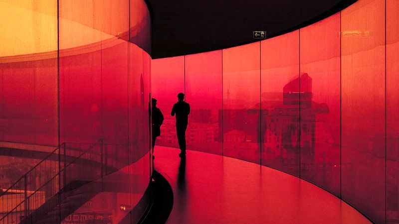 Long curving glass walkway looking out on a city. Image has a deep red tint and high contrast