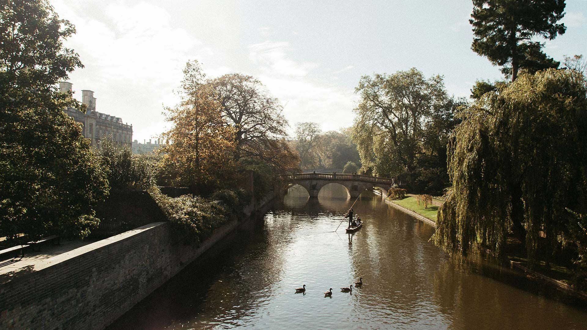 Hazy afternoon photo of river and bridge in Cambridge. Punt boat in middle-distance. Trees either side.