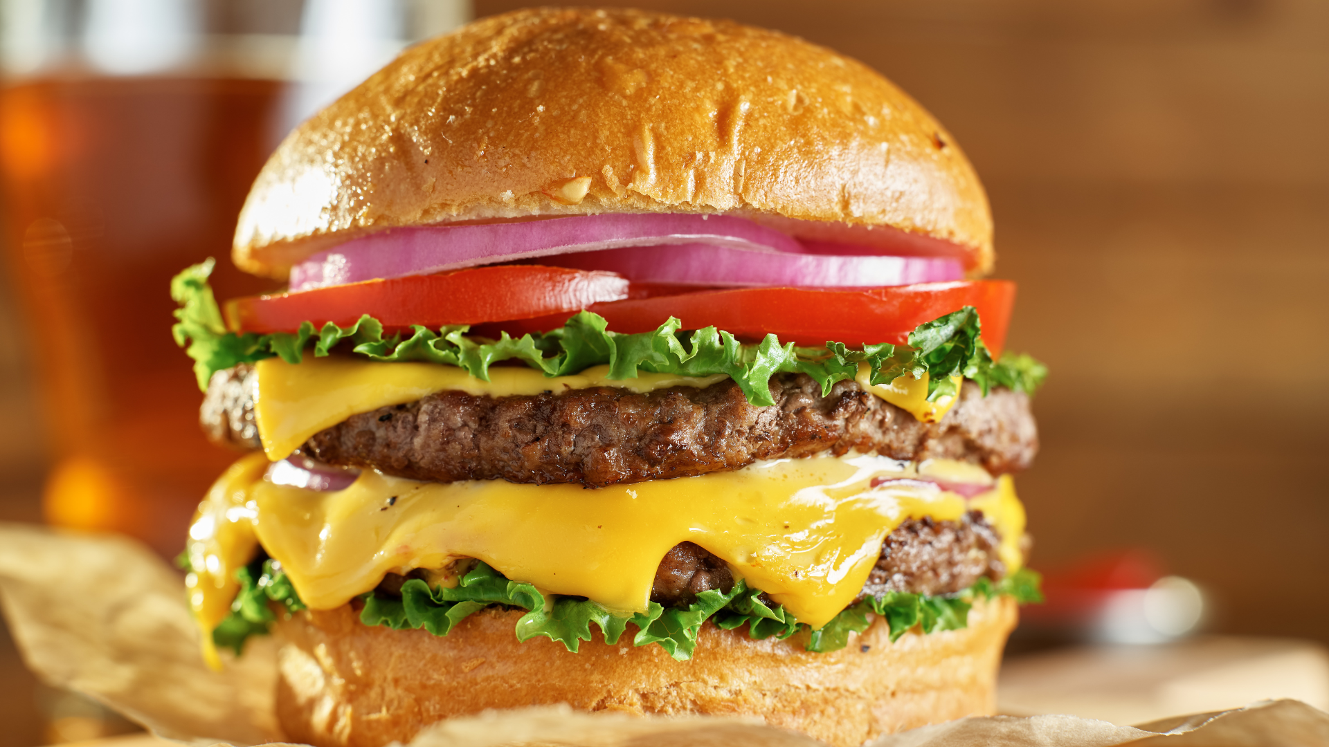 A double cheeseburger with lettuce , tomato , onions and a glass of beer in the background.