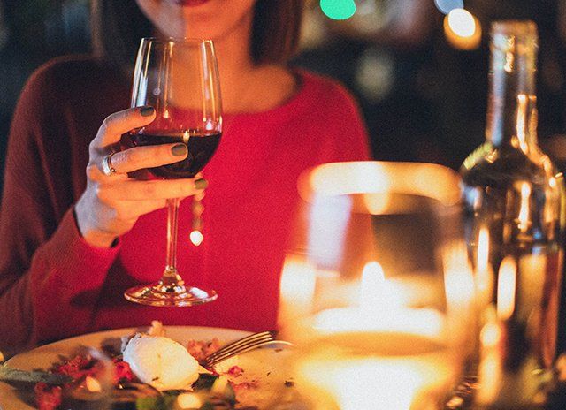 Woman in red sweater sips red wine at a dimly lit restaurant. Candle and food visible on the table.