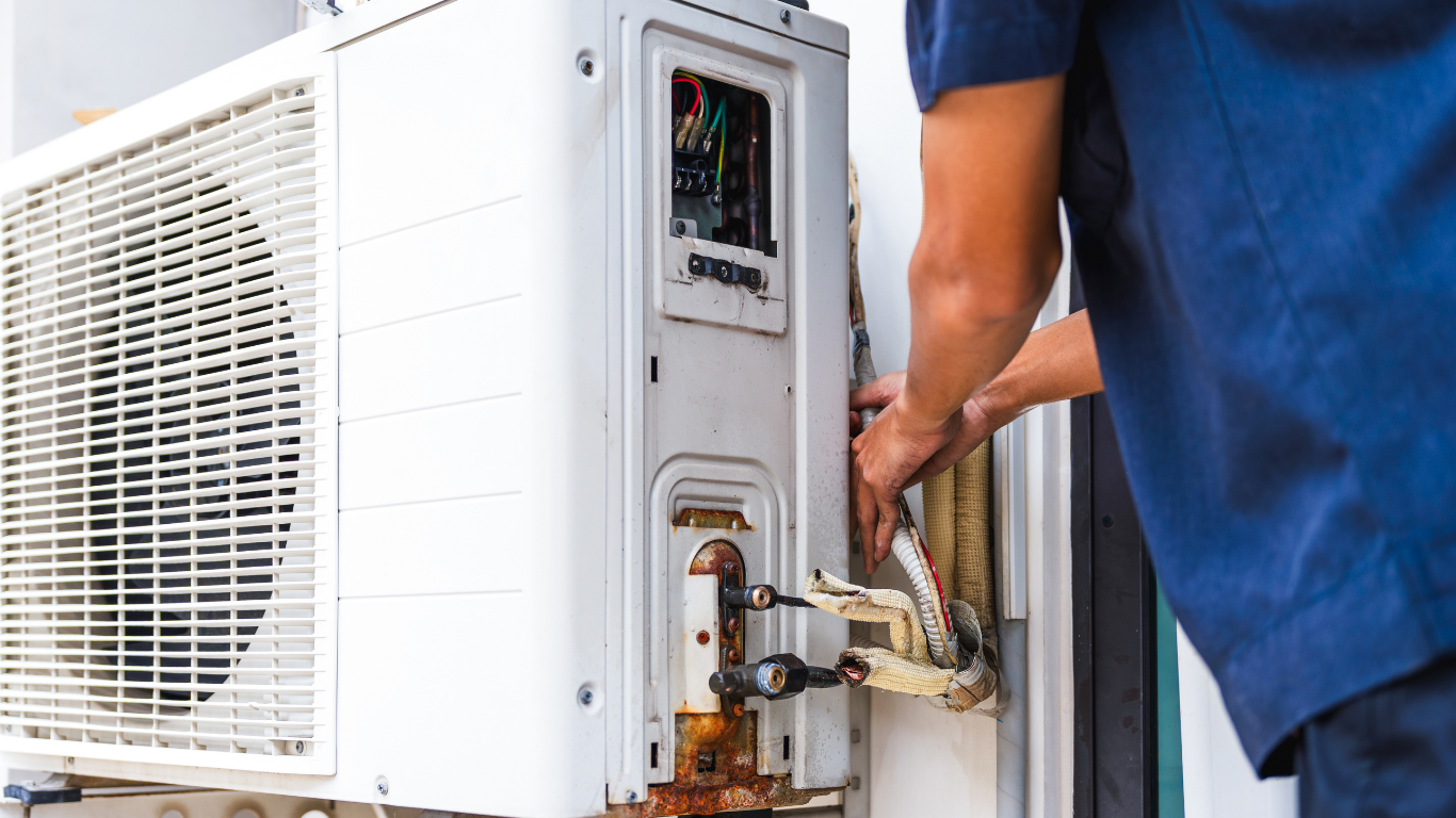 Technician repairing a white outdoor air conditioner unit with exposed wiring and rusted components