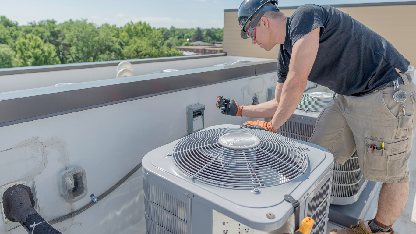 Technician kneeling on a rooftop beside an outdoor HVAC unit, inspecting it under bright sunlight