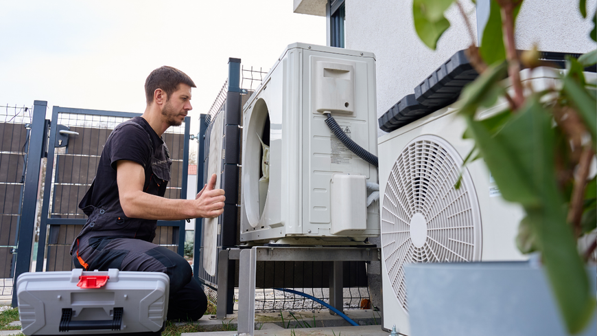 Technician inspecting outdoor HVAC unit beside a white building, with tools and equipment nearby.