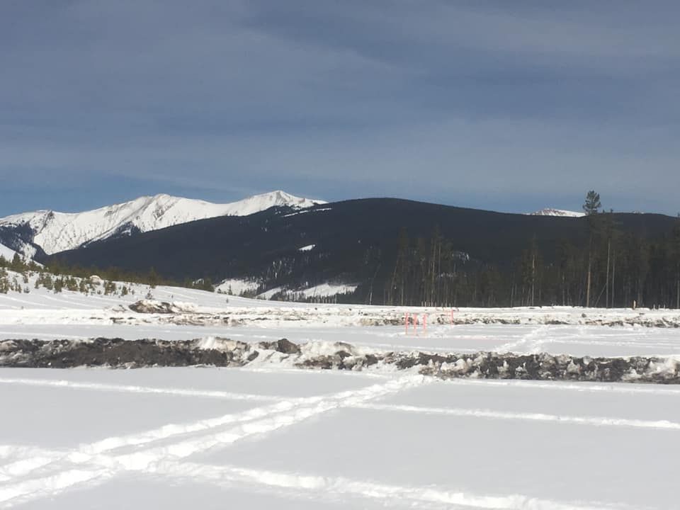 Snow-covered field with grid patterns and mounds of dirt, set against a backdrop of distant, snow-capped mountains.
